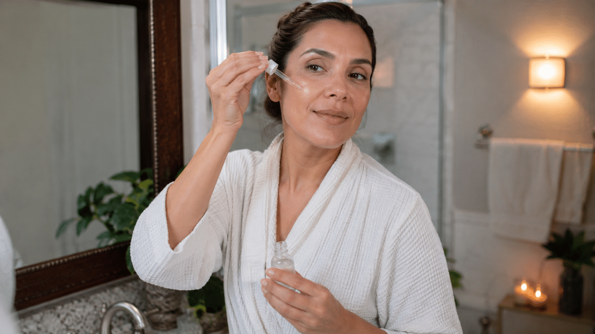 Mulher madura aplicando sérum transparente no rosto com conta-gotas, usando roupão branco em banheiro iluminado