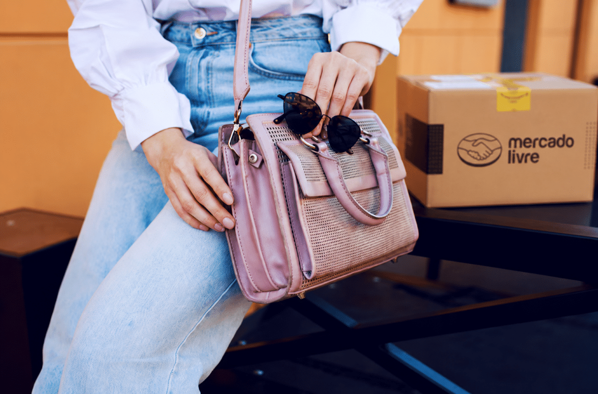 Mulher segurando bolsa rosa e óculos escuros próxima à caixa Mercado Livre em ambiente externo