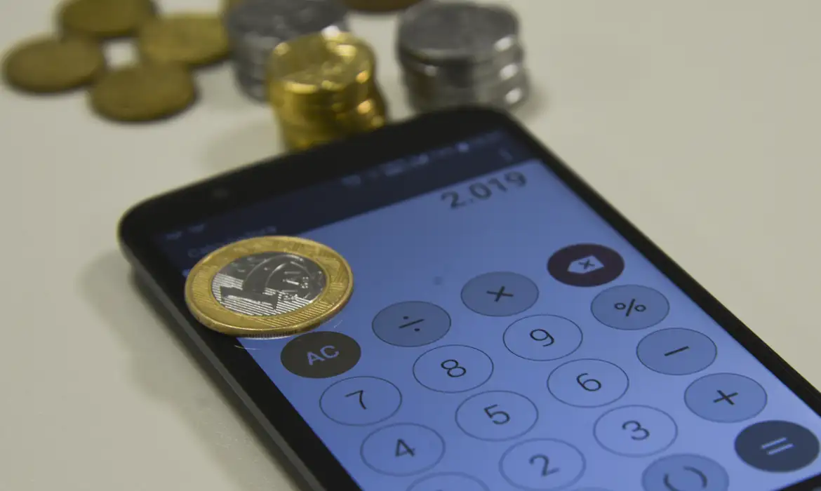Refis 2026: veja como o parcelamento 100% digital pode eliminar juros e multas 2 Moeda de 1 real sobre tela de smartphone com app de calculadora e outras moedas ao fundo