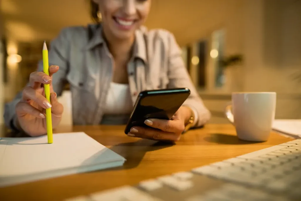 Nem toda compra internacional paga imposto! Veja os produtos que podem ficar livres da cobrança 2 Mulher sorridente utilizando celular e segurando um lápis sobre uma mesa com papéis, representando a consulta às regras de isenção de impostos em compras internacionais.